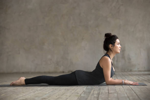 Young woman doing Sphinx exercise Young woman practicing yoga, doing Ardha bhudjangasana, Sphinx exercise, baby Cobra pose, working out, wearing sportswear, black pants and top, indoor full length, gray wall in yoga studio hugging knees stock pictures, royalty-free photos & images