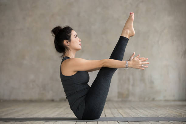 Young woman in boat pose Young yogi woman practicing yoga, doing Paripurna Navasana exercise, boat pose, working out, wearing sportswear, black pants and top, indoor full length, gray wall in yoga studio pilates photos stock pictures, royalty-free photos & images