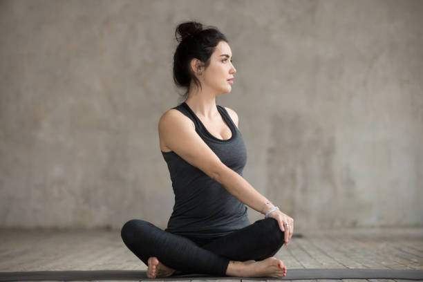 Young woman in Parivrtta Sukhasana pose Young sporty woman practicing yoga, doing Revolved Easy exercise, Parivrtta Sukhasana pose, working out, wearing sportswear, black pants and top, indoor full length, gray wall, yoga studio sukhasana stock pictures, royalty-free photos & images