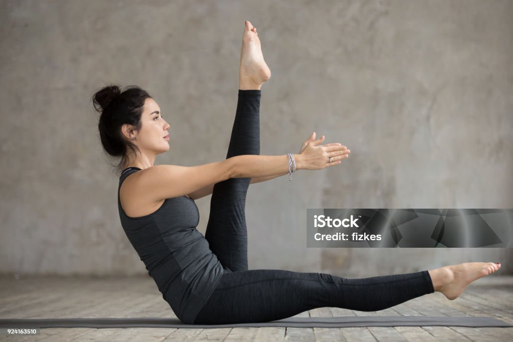 Young woman doing alternate leg stretch exercise Young sporty woman practicing, doing alternate leg stretch exercise, working out, wearing sportswear, black pants and top, indoor full length, gray wall in yoga studio Abdominal Muscle Stock Photo