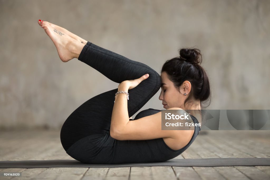 Young woman doing Knees to Chest exercise Young sporty woman practicing yoga, doing Knees to Chest exercise, Apanasana pose, working out, wearing sportswear, black pants and top, indoor full length, gray wall in yoga studio Pilates Stock Photo