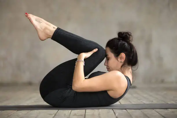 Young sporty woman practicing yoga, doing Knees to Chest exercise, Apanasana pose, working out, wearing sportswear, black pants and top, indoor full length, gray wall in yoga studio
