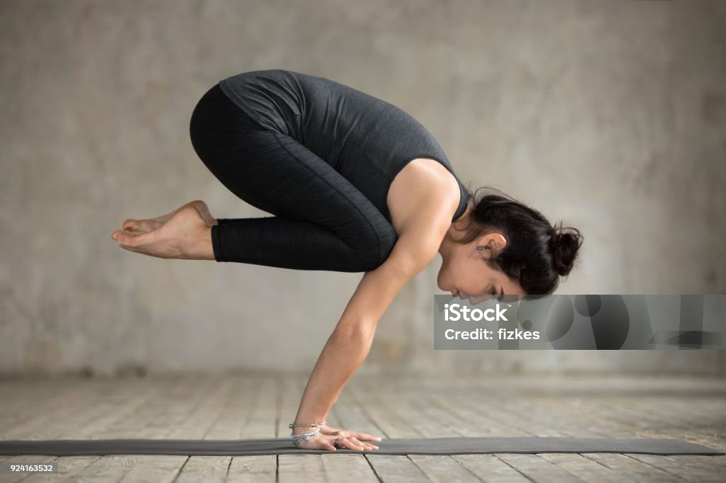 Young woman doing Bakasana exercise Young woman practicing yoga, doing Crane exercise, Bakasana pose, working out, wearing sportswear, black pants and top, indoor full length, gray wall in yoga studio Yoga Stock Photo