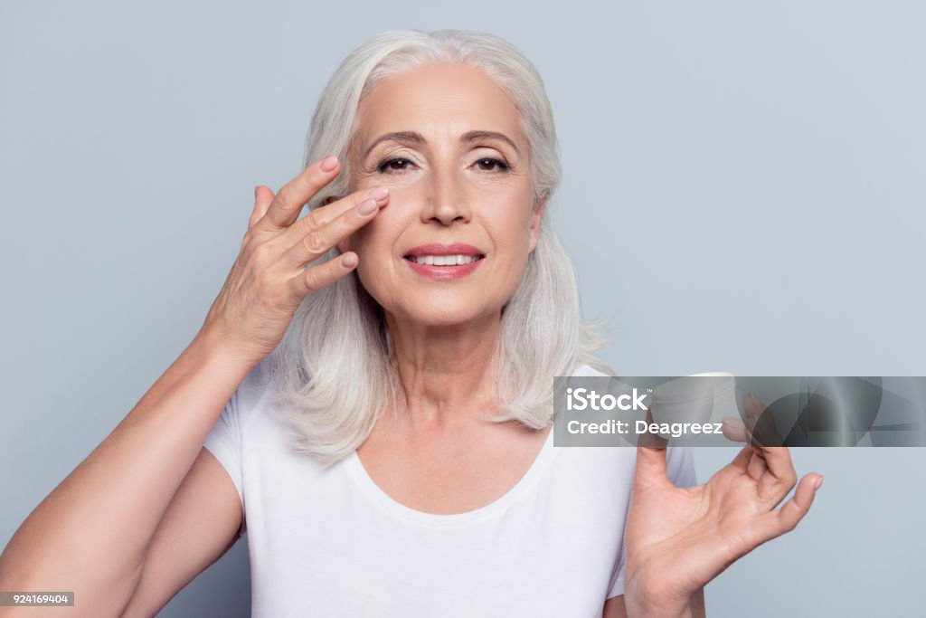 完璧なかなり、女性の目のクリームを適用する、灰色の背景の上のカメラを見て化粧品の瓶�を保持しています。 - 保湿クリームのロイヤリティフリーストックフォト
