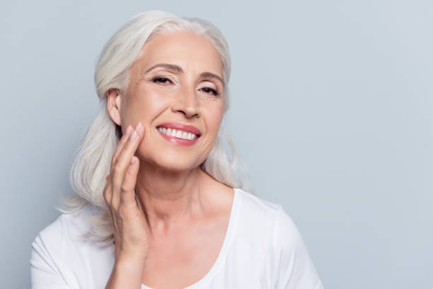 encantador, bastante, mujer tocar su suave rostro perfecto de la piel con los dedos, sonriendo a la cámara sobre fondo gris, uso de día, crema de noche facial, procedimientos de cosmetología - wrinkle treatment fotografías e imágenes de stock