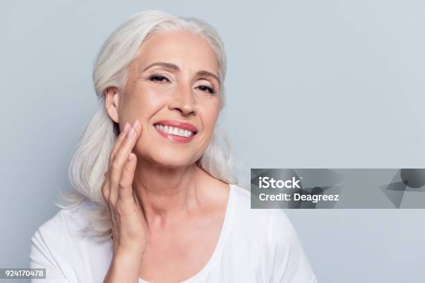 Foto de Charmoso Bonito Velha Mulher Tocando Seu Rosto Suave Perfeito Da Pele Com Os Dedos Sorrindo Para A Câmera Sobre Um Fundo Cinza Usando De Dia Creme De Noite Cara Procedimentos De Cosmetologia e mais fotos de stock de Face Humana