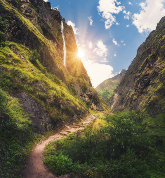 Amazing Himalayan mountains covered green grass, high waterfall, beautiful path, green trees, blue sky with yellow sun and clouds in Nepal at sunset. Mountain canyon. Travel in Himalayas.Landscape