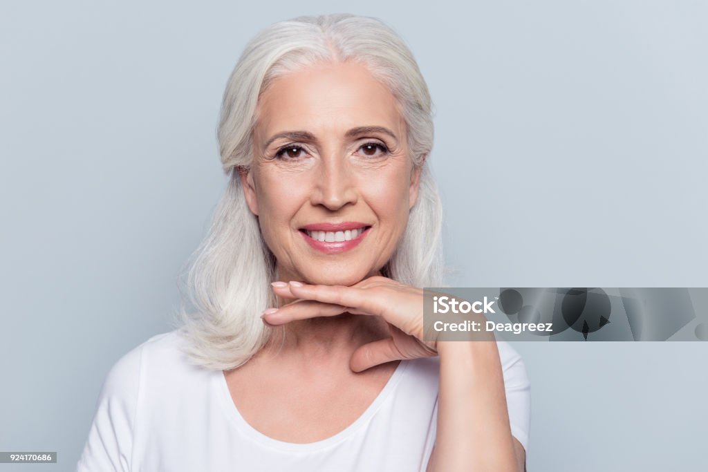 Charmante vieille femme tenant la main sous le menton avec sourire rayonnant, regardant la caméra sur fond gris - Photo de Femmes seniors libre de droits
