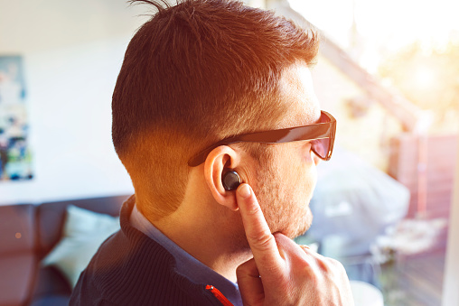 Truely wireless earbuds placed in the ear of a man. The earbuds are connected to a mobile phone and can be controlled by tapping the device. It plays music or a podcast. The earbuds have built in microphones and handle phone calls hands-free. The earbuds also work as a digital assistant, and the user can ask questions and get answers.