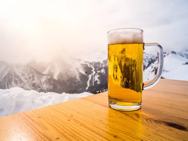 Photo of Glass or pint of a beer on a wood table on the top of the alps. Sun light is shining on the winter landscape.