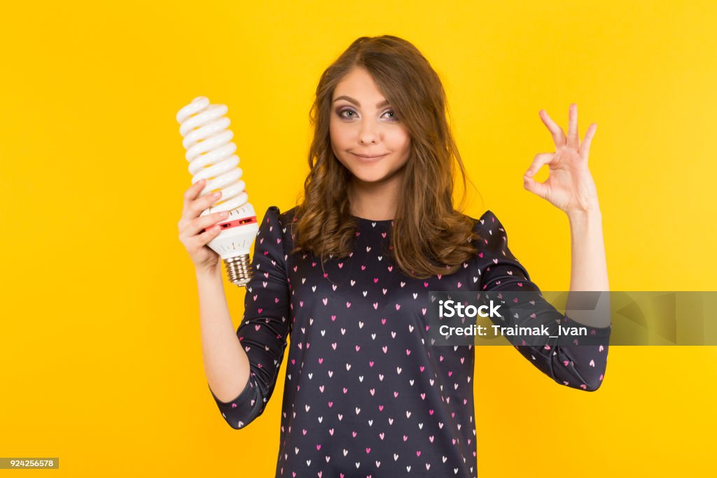 Attractive woman with energy saving bulb Portrait of attractive brunette woman holding spiral light bulb and showing OK sign isolated on yellow background advertising energy saving concept. Light Bulb Stock Photo