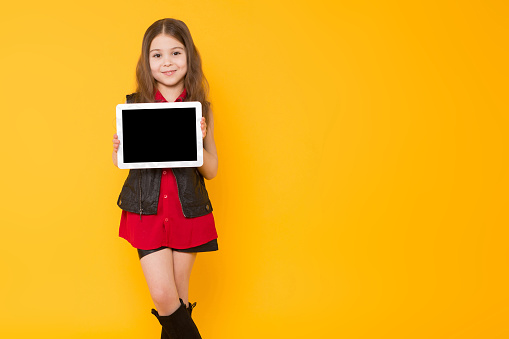 Portrait of little cute girl in red shirt and high boots presenting digital tablet pc isolated on orange background advertising network concept.