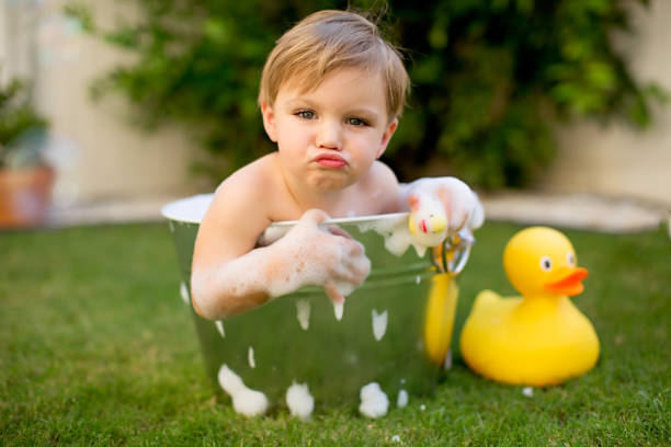 Little boy is in a bucket with bubbles in a hot summer day. He is in the backyard and playing with rubber duckies.