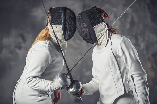 Female fencers in protective clothing are fighting on dark gray background