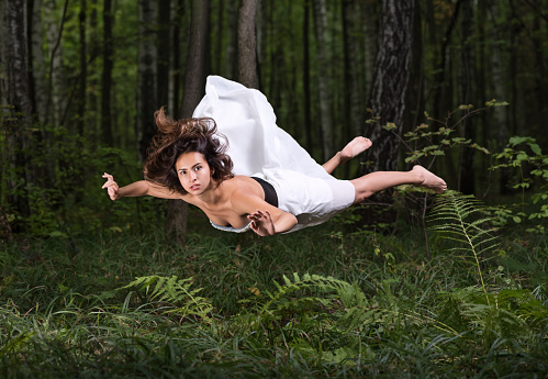 Zero gravity. Young beautiful woman flying in a dream in a summer forest. White dress and hair in the air. Surprise and light fright