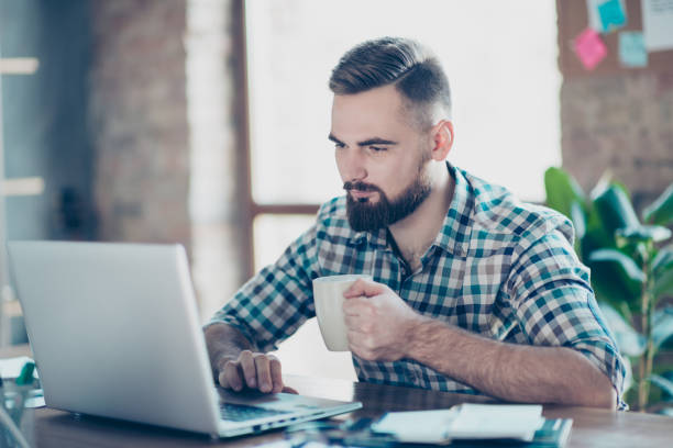 Concept of having a break at work. Concentrated calm peaceful serious office worker  watching carefully a video on the internet using his laptop and drinking tea