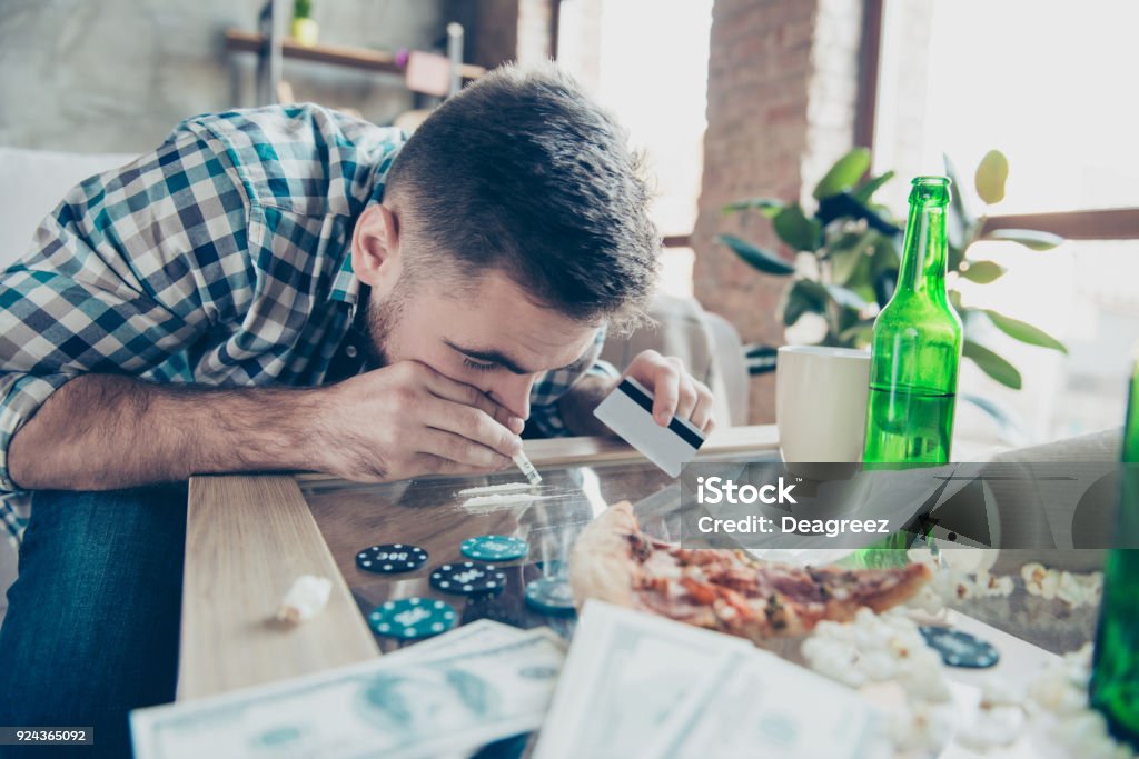 Close up portrait of exhausted drunk overdosed guy dressed in checkered shirt, he is sniffing a snorting line made of drugs on a table Credit Card Stock Photo