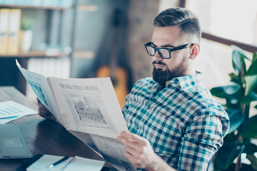 Careless concentrated clever bearded guy clothed in casual checkered shirt is reading a newspaper at work, he has a break, he is sitting at the table