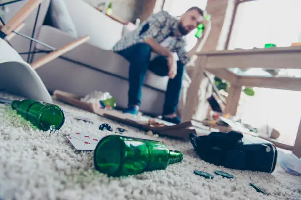 Concept of having headache after global party. Close up low-angle photo of empty green transparent beer bottles lying on nappy beige carpet, guy with nausea sitting on sofa in ob blurred background