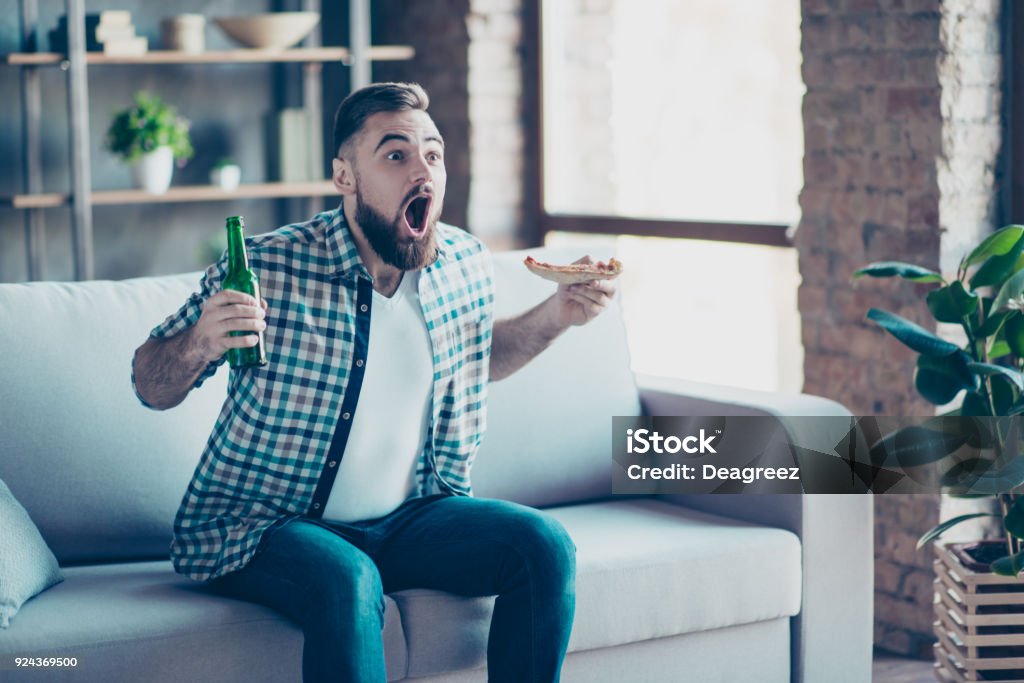 OMG ! Objectif ! Funny heureux excité étonné surpris fou habillé en jeans et chemise à carreaux, il mange pizza et boire de la bière, assis sur un canapé et regarder la finale du jeu de sport - Photo de Télévision libre de droits