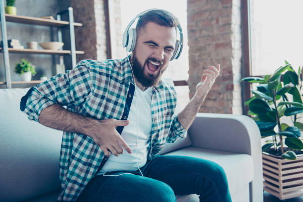 attrayant, barbu homme assis sur le canapé dans le salon, avoir un casque sur la tête, écoute sa musique préférée, chanter une chanson rêver comme jouer de la guitare - bachelor home photos et images de collection