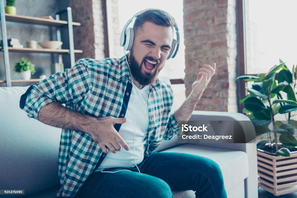 Attractive, bearded man sitting on the couch in living room, having headphones on his head, listening his favorite music, singing a song dreaming like playing guitar Music Stock Photo