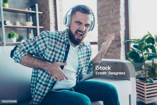 Attractive Bearded Man Sitting On The Couch In Living Room Having Headphones On His Head Listening His Favorite Music Singing A Song Dreaming Like Playing Guitar Stock Photo - Download Image Now