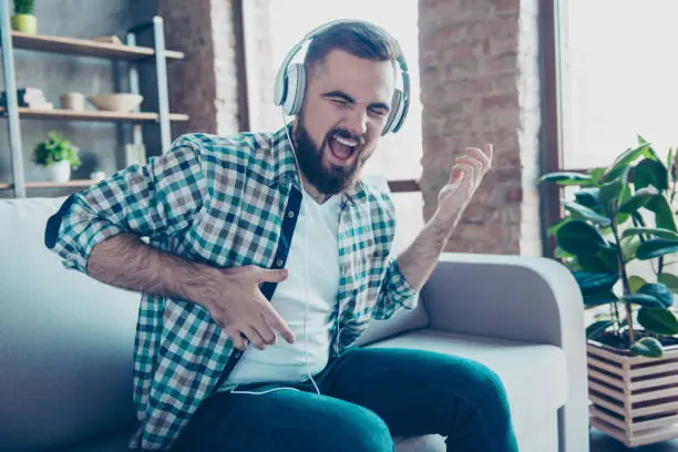 Photo of Attractive, bearded man sitting on the couch in living room, having headphones on his head, listening his favorite music, singing a song dreaming like playing guitar