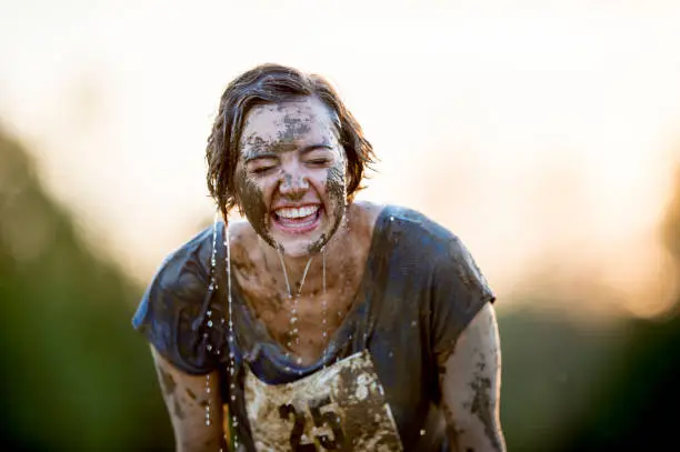 A Caucasian woman is standing outdoors. She has just completed a mud run. She is puring water on her face to clean off.