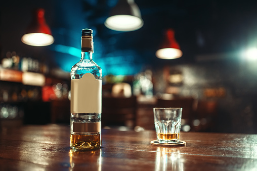 Bottle of strong alcohol and glass on bar counter, closeup. Restaurant interior, nobody, blur effect