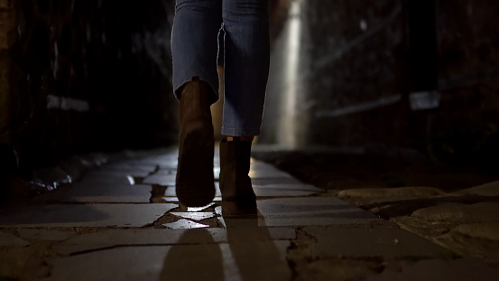 Woman feet in boot walking on stone pavement in old alley dark street