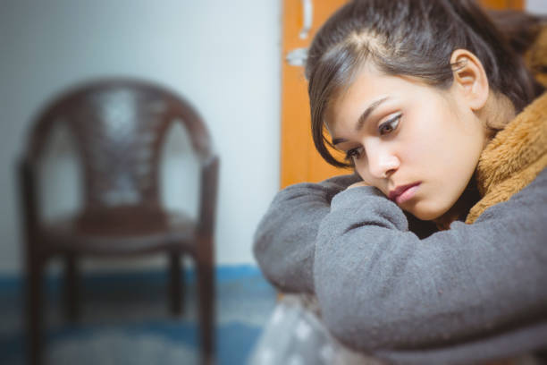 Asian, Indian sad, serene, single young woman at home. She is looking down, resting her head on hands thinking deeply with blank expression. There is empty chair is placed in the background.