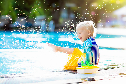 Baby with toy boat in swimming pool. Little boy learning to swim in outdoor pool of tropical resort. Swimming with kids. Healthy sport activity for children. Sun protection swim wear. Water toys.