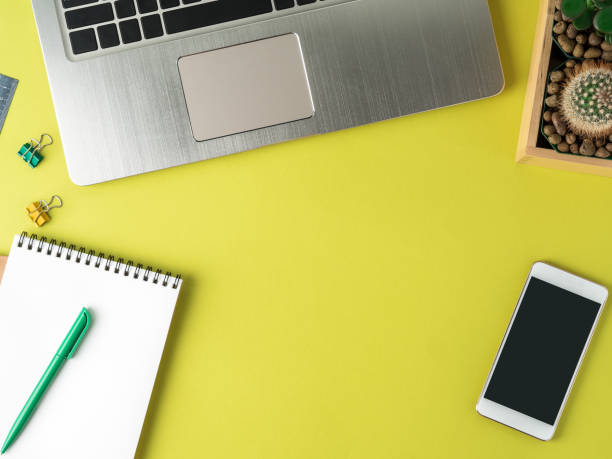 istock Top view of modern bright yellow office desktop with blank notepad, computer, smartphone, cactus, succulent. Mock up, empty space 924494400