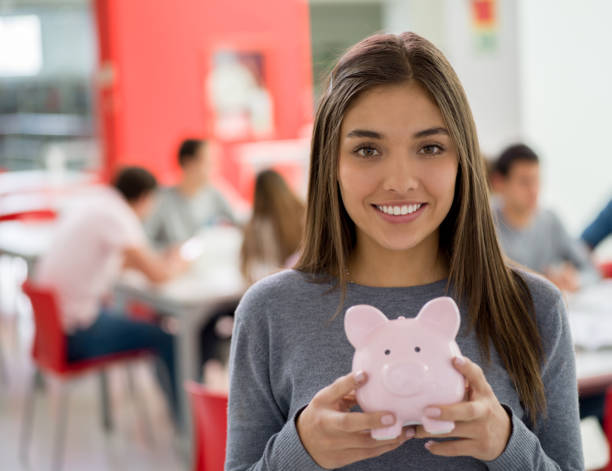 schöne studentin in der bibliothek hält ein sparschwein, blick auf die kamera zu lächeln - student loans stock-fotos und bilder