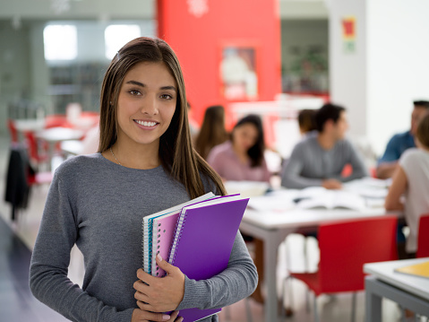 Beautiful young female student looking at camera smiling very happy at the library while holding her notebooks