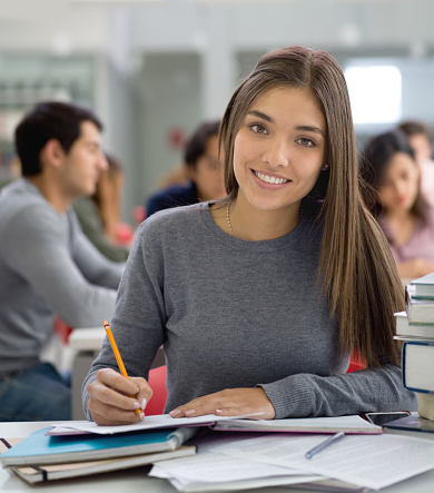 Female student at the library studying for an exam using books and taking notes looking at camera smiling while other sudents are studying at background