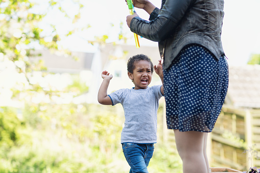 afro american toddler boy, 3 years old, screaming ad crying, diappointed about something his mother will not do. Mother is there too, but focus is on the boy. They are outdoors in backyard, it is summer