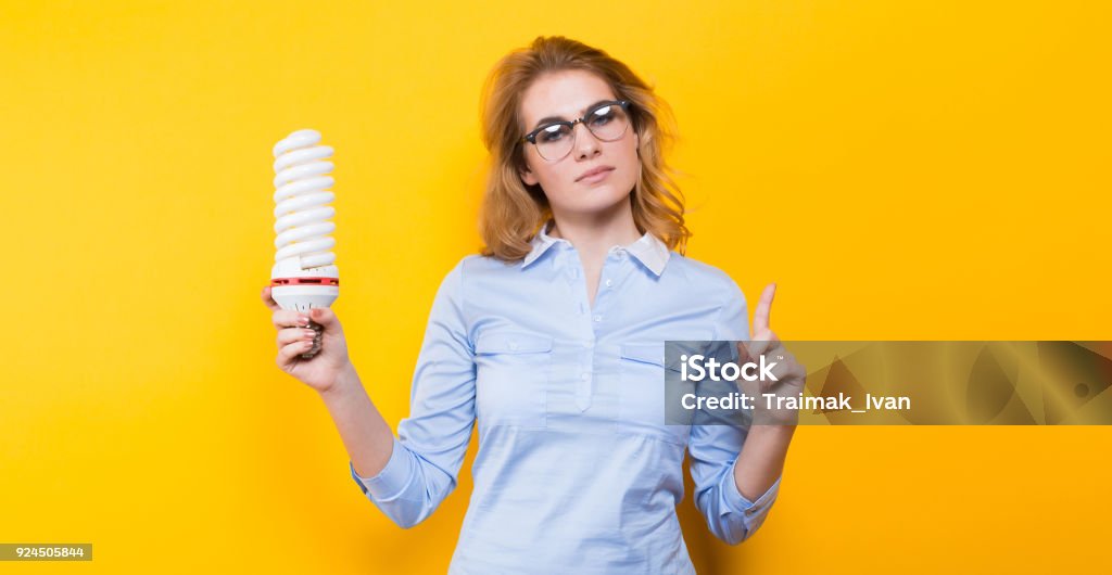 Attractive woman with energy saving bulb Portrait of attractive blonde woman in blue shirt and eyeglasses holding spiral light bulb and her finger up isolated on yellow background advertising energy saving concept. Adult Stock Photo