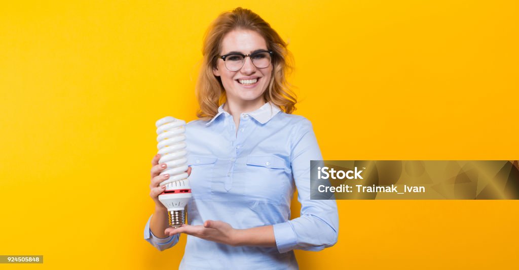 Attractive woman with energy saving bulb Portrait of attractive blonde woman in blue shirt and eyeglasses holding spiral light bulb a isolated on yellow background advertising energy saving concept. Adult Stock Photo