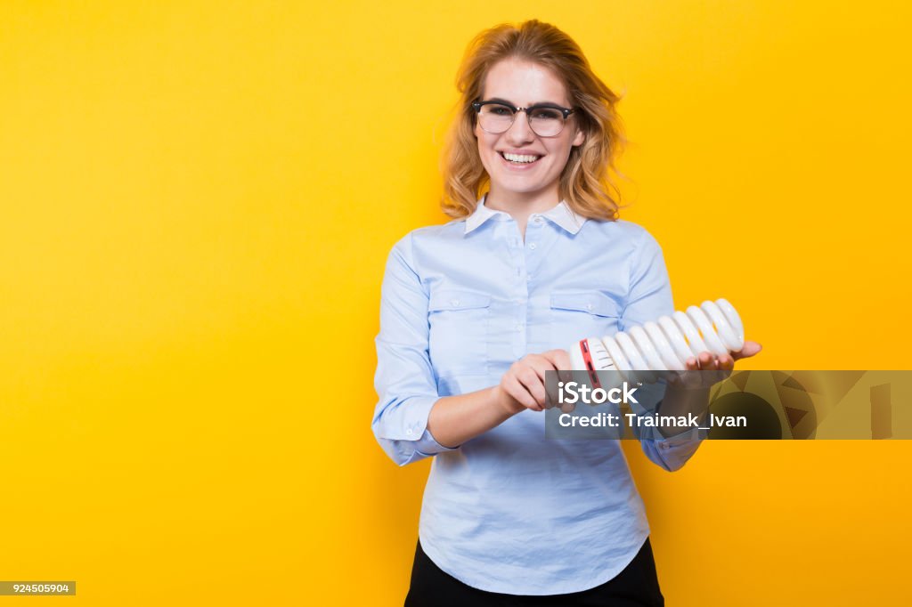 Attractive woman with energy saving bulb Portrait of attractive blonde woman in blue shirt and eyeglasses holding spiral light bulb isolated on yellow background advertising energy saving concept. Adult Stock Photo