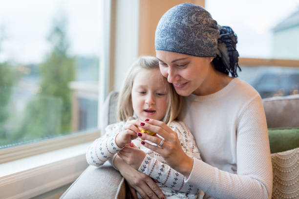 Young mom with cancer holds her daughter A beautiful young woman with cancer holds her preschool-age daughter in her lap by their living window. They are holding and playing with a little plastic toy and mom is smiling. She is also wearing a headscarf. cancer illness stock pictures, royalty-free photos & images