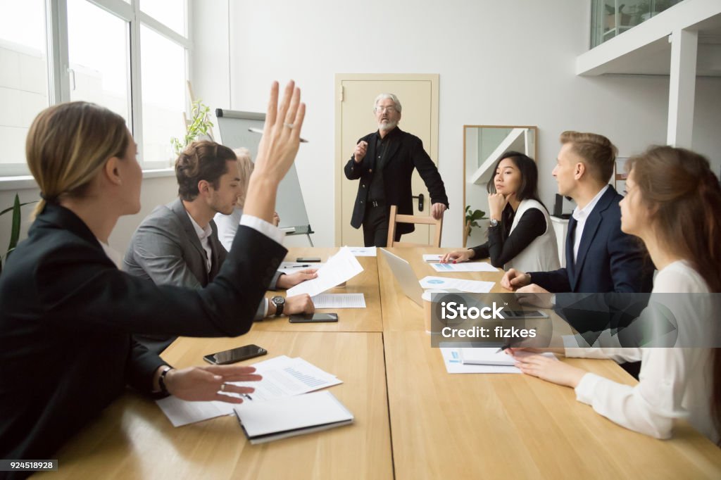Businesswoman raising hand asking senior coach questions at team meeting Businesswoman raising hand up at diverse team meeting sitting at conference table, student asking senior teacher mentor coach questions during seminar lecture training, corporate education concept Asking Stock Photo