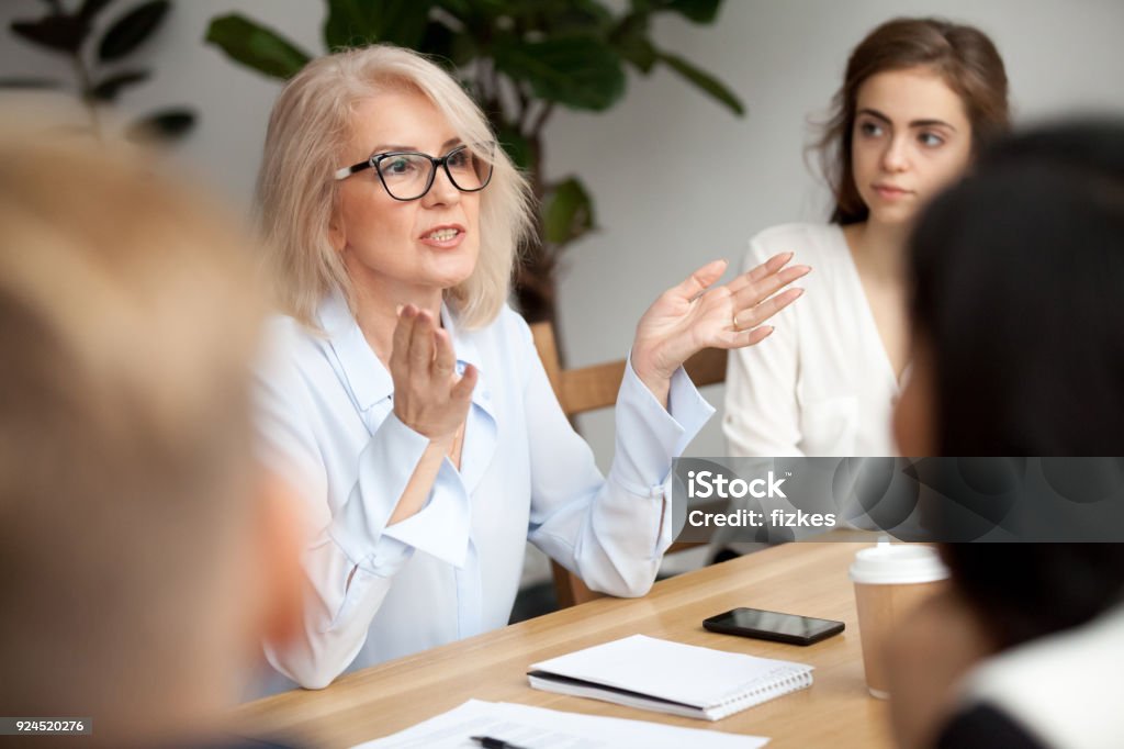Aged businesswoman, teacher or business coach speaking to young people Attractive aged businesswoman, teacher or mentor coach speaking to young people, senior woman in glasses teaching audience at training seminar, female business leader speaker talking at meeting Women Stock Photo
