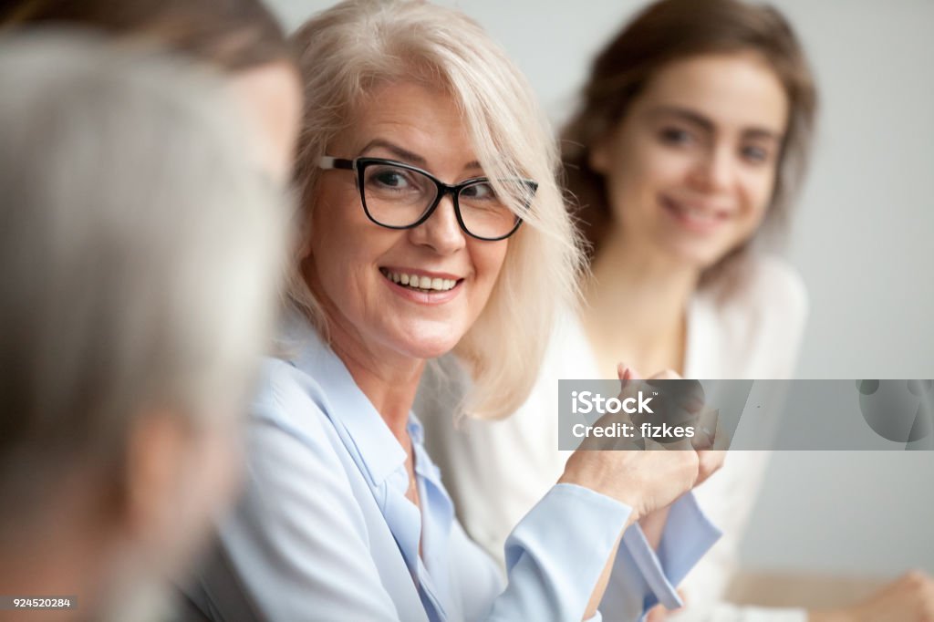 Smiling aged businesswoman looking listening to colleague at team meeting Smiling aged businesswoman in glasses looking at colleague at team meeting, happy attentive female team leader listening to new project idea, coach mentor teacher excited by interesting discussion Senior Adult Stock Photo