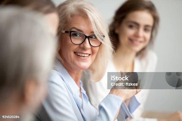 Smiling Aged Businesswoman Looking Listening To Colleague At Team Meeting Stock Photo - Download Image Now