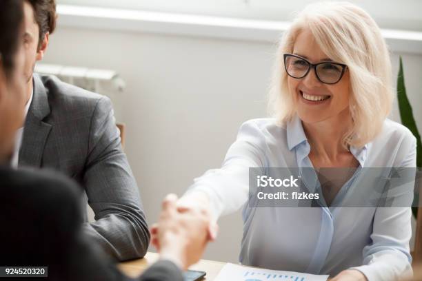 Smiling Attractive Mature Businesswoman Handshaking Businessman At Group Meeting Negotiations Stock Photo - Download Image Now