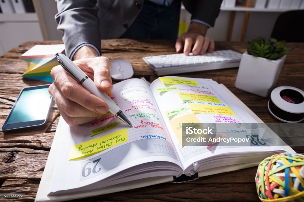 Businessperson's Hand Writing Schedule In Diary With Pen Close-up Of A Businessperson's Hand Writing Schedule In Diary With Pen On Wooden Desk Personal Organizer Stock Photo