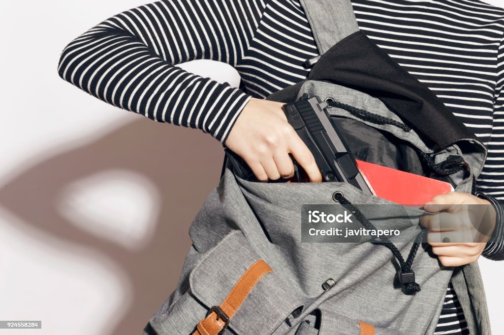 Girl hiding a gun in her school backpack. Girl hiding a gun in her school backpack. Concealed carry weapon for protection themselves concept. Indoors, over a white wall. Gun Stock Photo