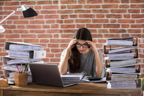 Exhausted Businesswoman Working At Office With Stack Of Folders On Desk